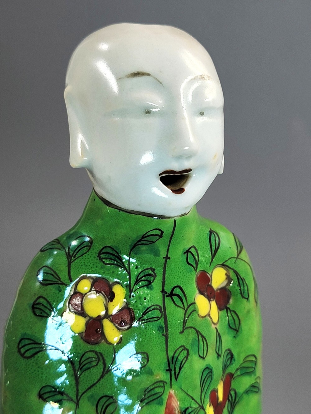 An antique Chinese ceramic incense holder depicting a smiling boy in a green floral jacket seated on a red base. View VII