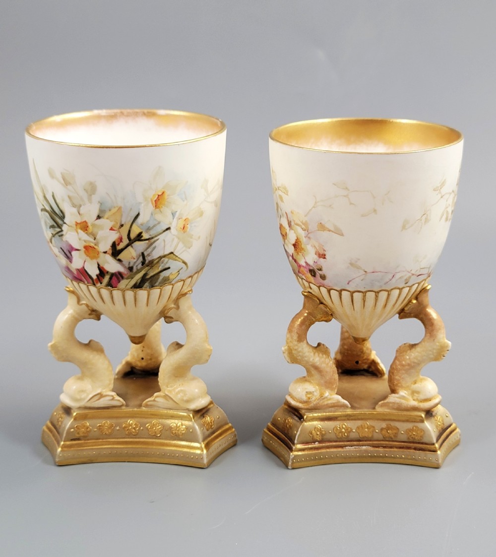 A pair of late Victorian Doulton Burslem porcelain beaker-form vases featuring hand-painted floral motifs and interior gilding, supported by three sculpted dolphin pedestals resting on moulded triangular gold bases. View II