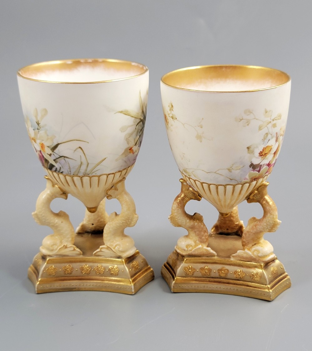 A pair of late Victorian Doulton Burslem porcelain beaker-form vases featuring hand-painted floral motifs and interior gilding, supported by three sculpted dolphin pedestals resting on moulded triangular gold bases. View V