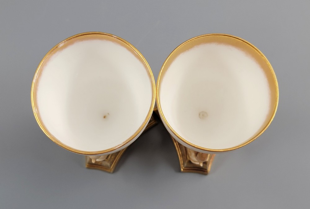 A pair of late Victorian Doulton Burslem porcelain beaker-form vases featuring hand-painted floral motifs and interior gilding, supported by three sculpted dolphin pedestals resting on moulded triangular gold bases. View VI