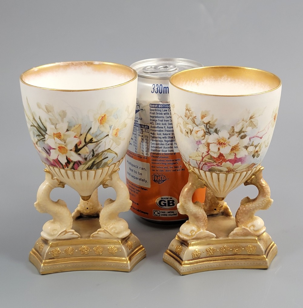 A pair of late Victorian Doulton Burslem porcelain beaker-form vases featuring hand-painted floral motifs and interior gilding, supported by three sculpted dolphin pedestals resting on moulded triangular gold bases. View X