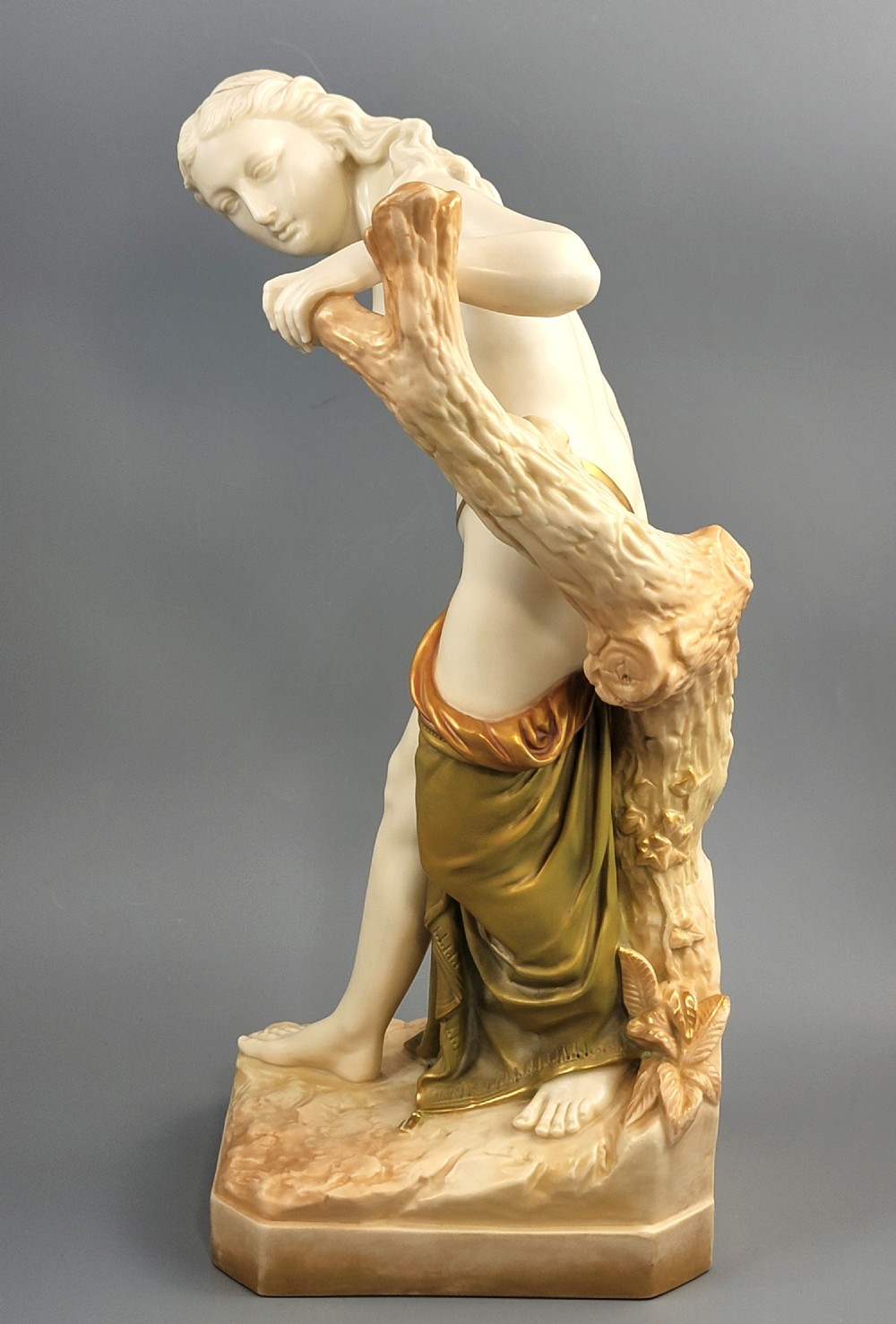 An Edwardian Royal Worcester porcelain figure depicting a classical female bather leaning against a tree trunk, rendered in cream porcelain with gold shot enamel drapery. View II