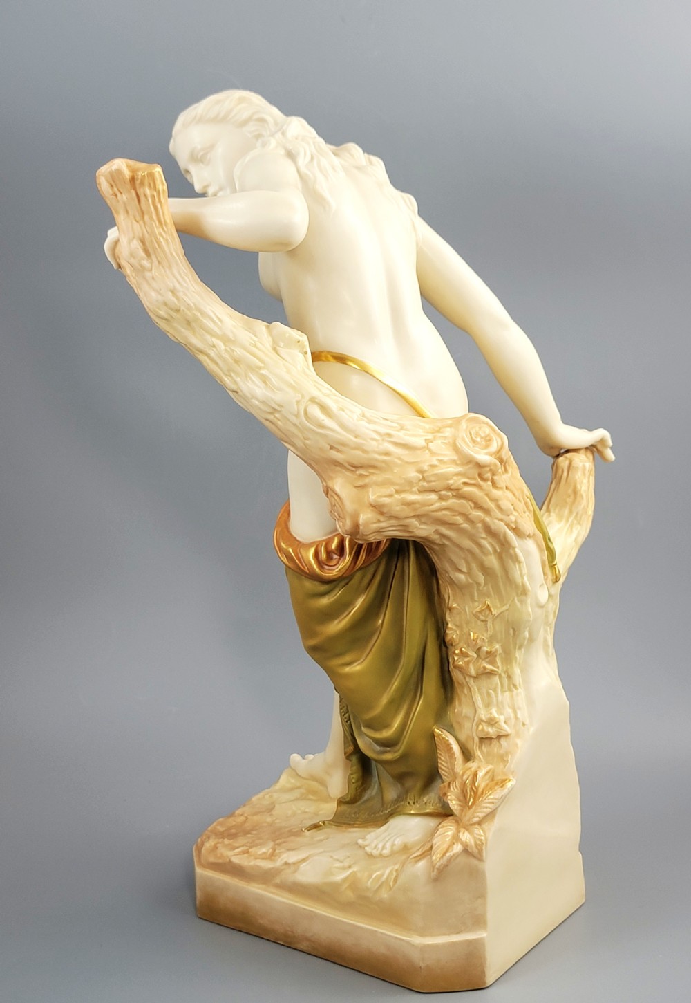 An Edwardian Royal Worcester porcelain figure depicting a classical female bather leaning against a tree trunk, rendered in cream porcelain with gold shot enamel drapery. View III