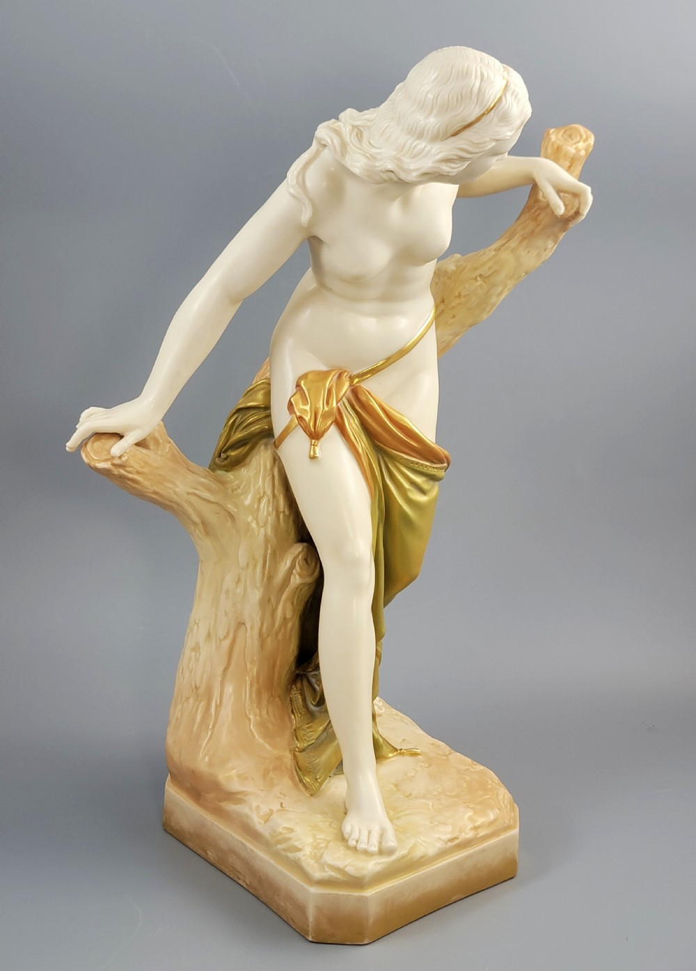An Edwardian Royal Worcester porcelain figure depicting a classical female bather leaning against a tree trunk, rendered in cream porcelain with gold shot enamel drapery. View VII