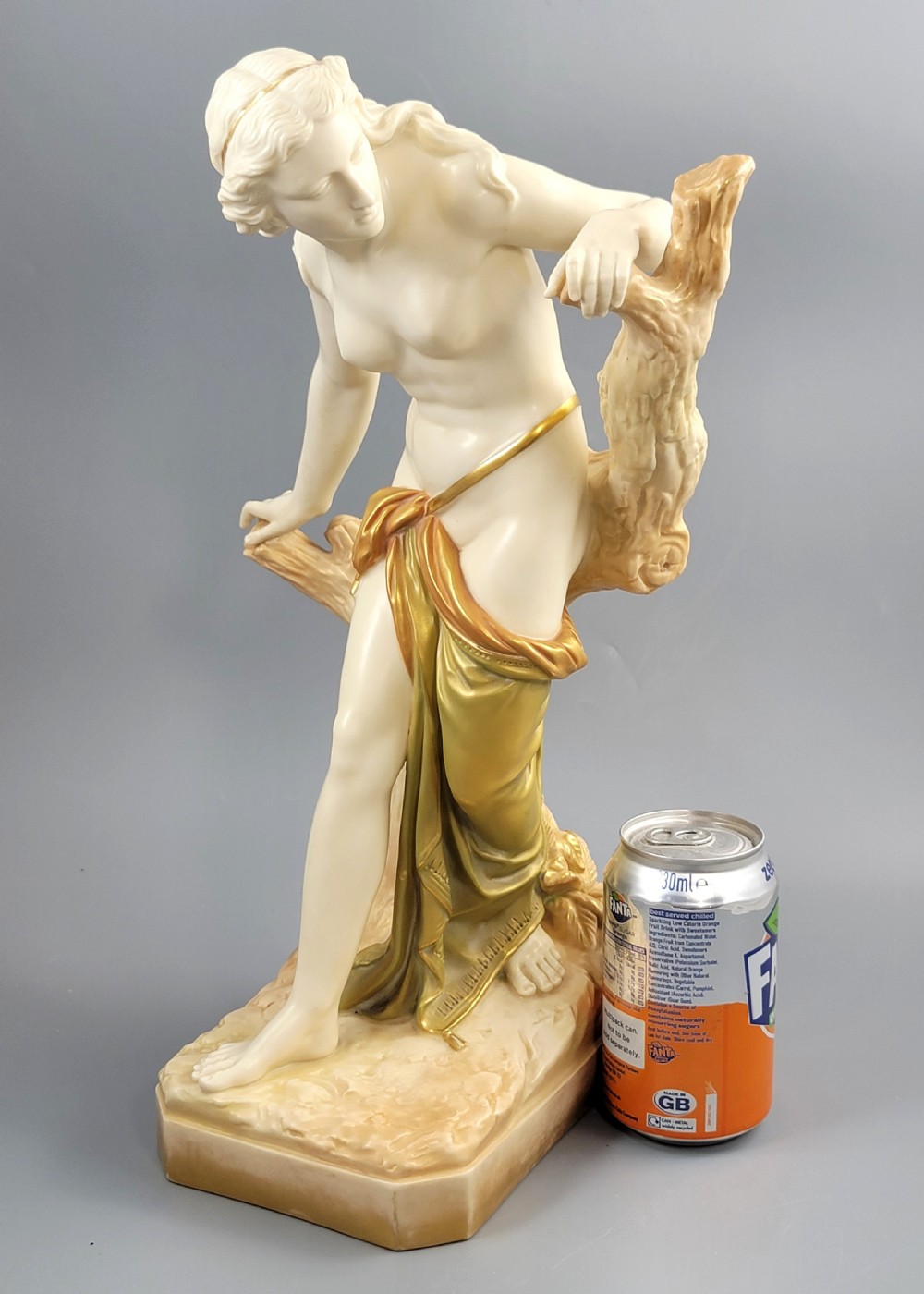 An Edwardian Royal Worcester porcelain figure depicting a classical female bather leaning against a tree trunk, rendered in cream porcelain with gold shot enamel drapery. View X