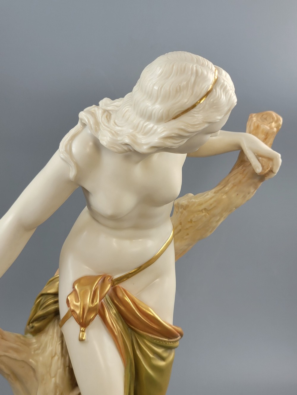 An Edwardian Royal Worcester porcelain figure depicting a classical female bather leaning against a tree trunk, rendered in cream porcelain with gold shot enamel drapery. View XII