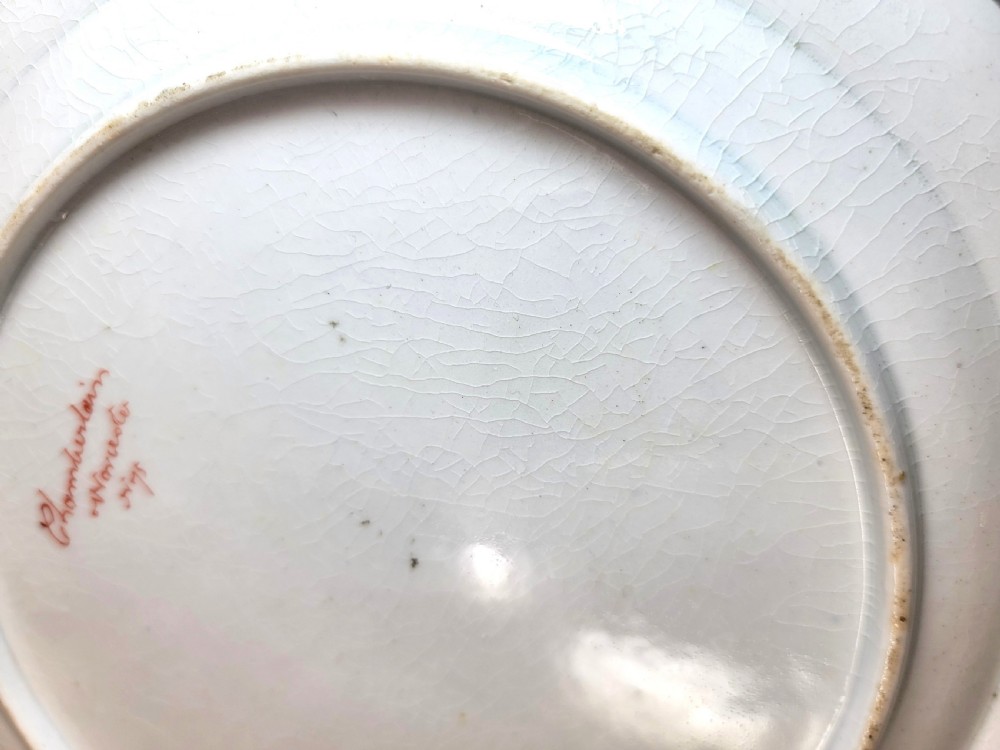 An antique circular porcelain dessert plate featuring hand-painted panels in red, green, and blue enamels surrounding a central family heraldic coat of arms. View V