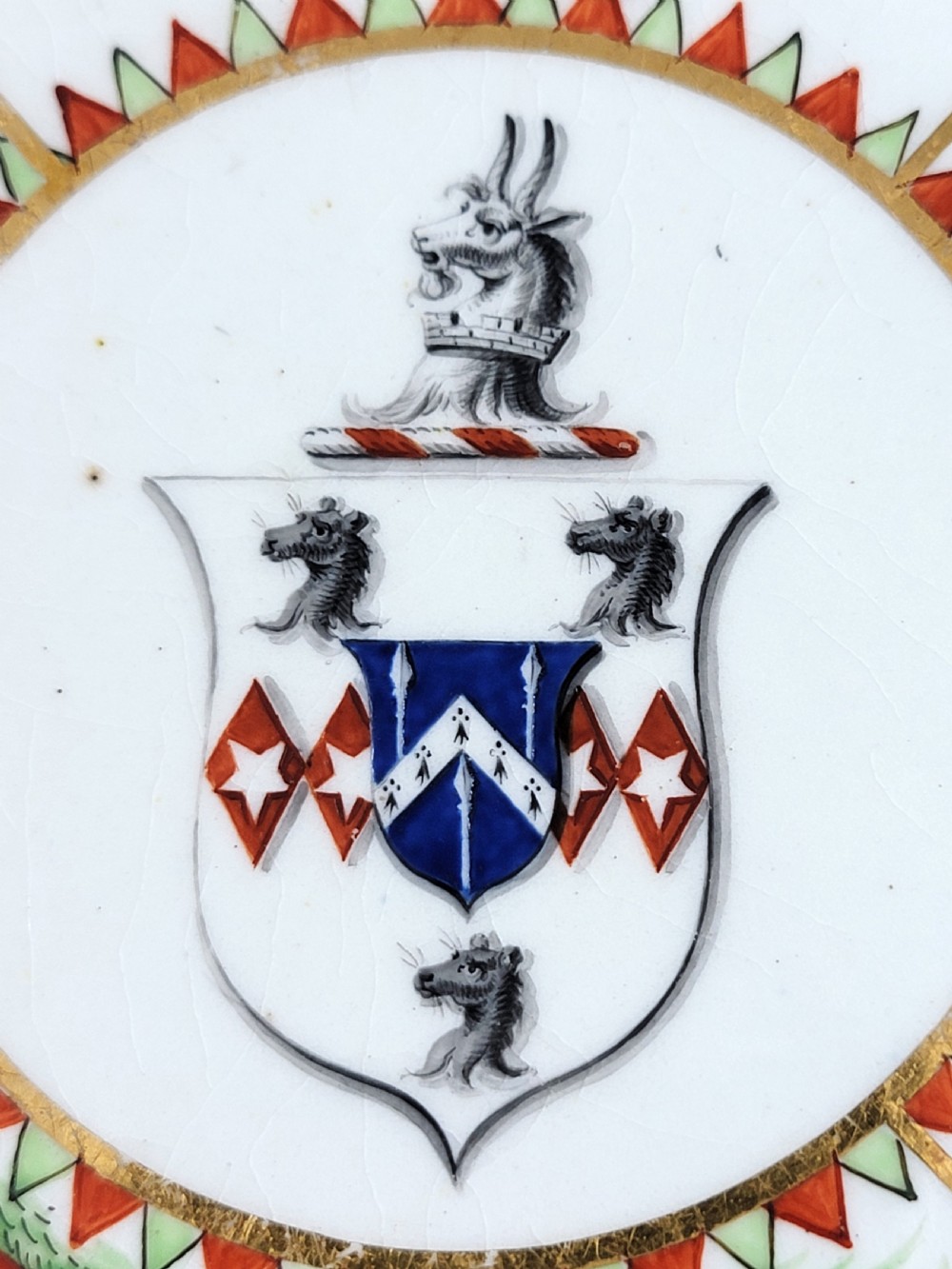An antique circular porcelain dessert plate featuring hand-painted panels in red, green, and blue enamels surrounding a central family heraldic coat of arms. View VI