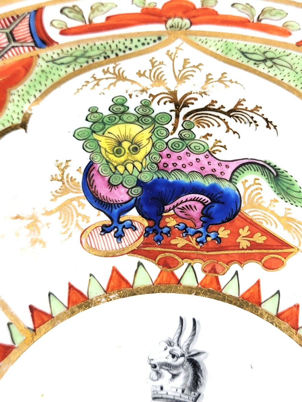 An antique circular porcelain dessert plate featuring hand-painted panels in red, green, and blue enamels surrounding a central family heraldic coat of arms. View VII