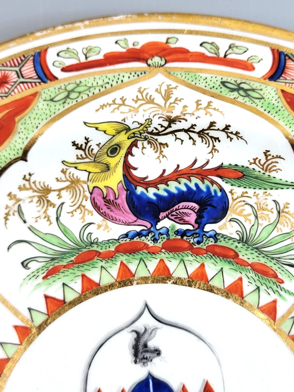 An antique circular porcelain dessert plate featuring hand-painted panels in red, green, and blue enamels surrounding a central family heraldic coat of arms. View IX
