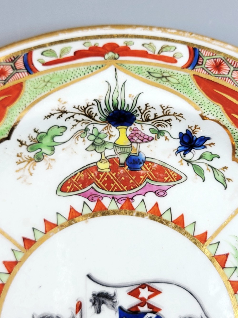 An antique circular porcelain dessert plate featuring hand-painted panels in red, green, and blue enamels surrounding a central family heraldic coat of arms. View X