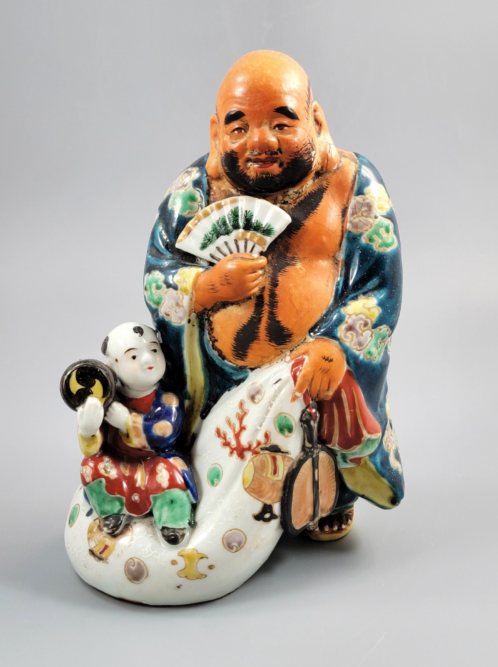 A Japanese Satsuma earthenware figure depicting Hotei in patterned blue robes holding a fan, standing next to a small child on a patterned sack.