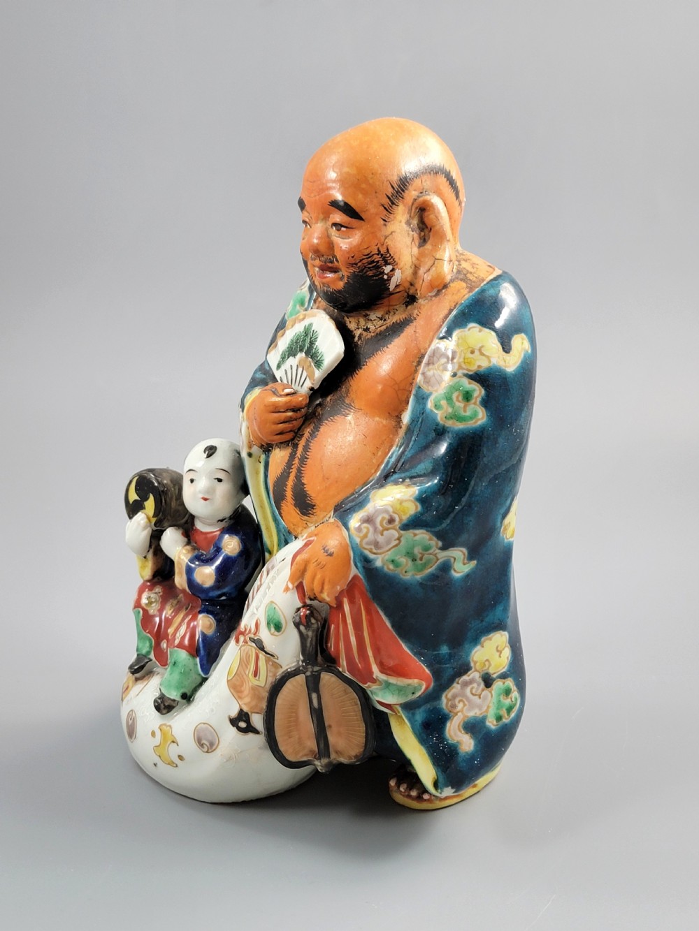 A Japanese Satsuma earthenware figure depicting Hotei in patterned blue robes holding a fan, standing next to a small child on a patterned sack. View II