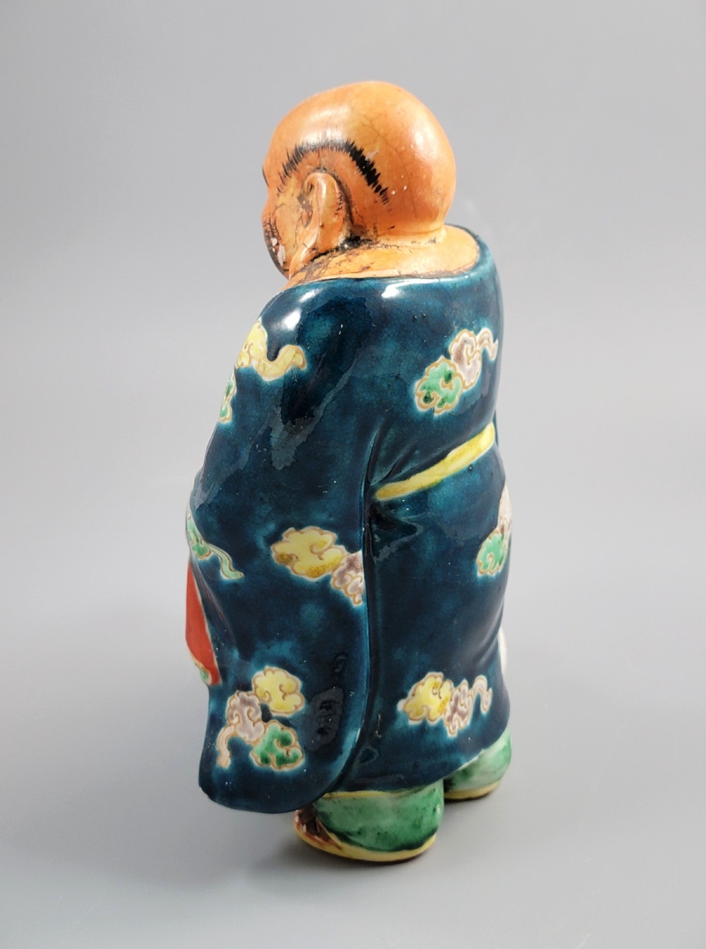 A Japanese Satsuma earthenware figure depicting Hotei in patterned blue robes holding a fan, standing next to a small child on a patterned sack. View III