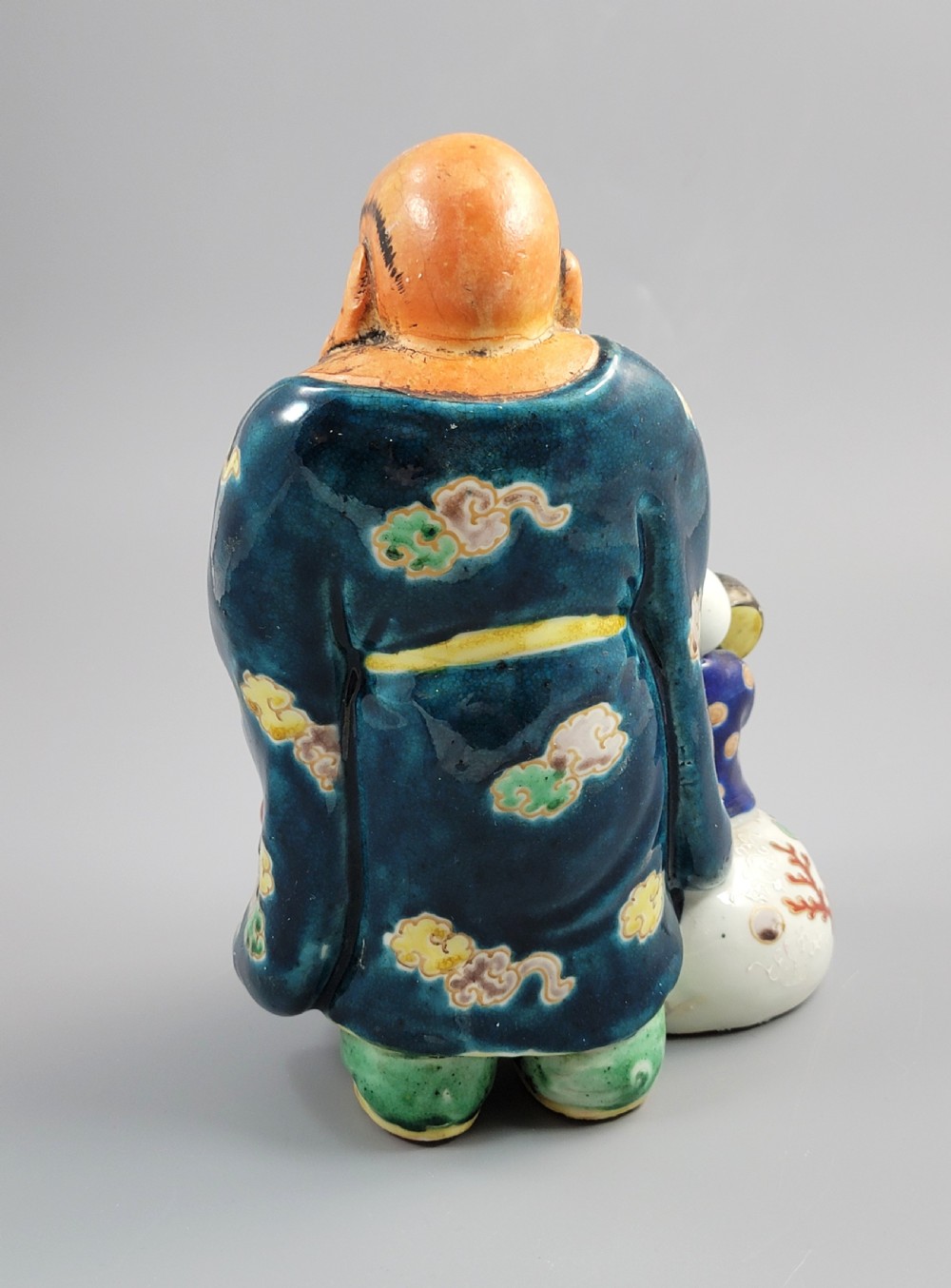 A Japanese Satsuma earthenware figure depicting Hotei in patterned blue robes holding a fan, standing next to a small child on a patterned sack. View IV