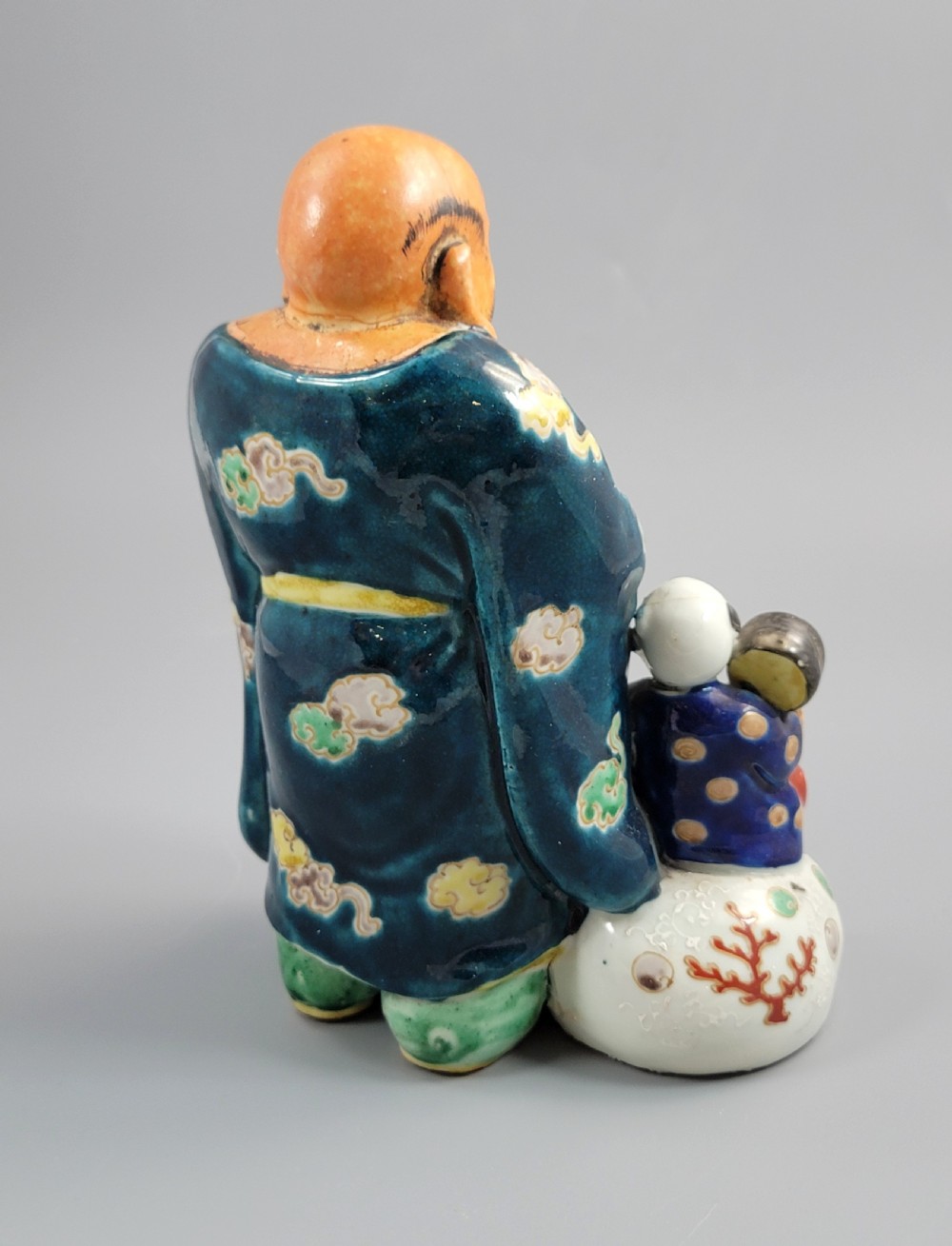 A Japanese Satsuma earthenware figure depicting Hotei in patterned blue robes holding a fan, standing next to a small child on a patterned sack. View V