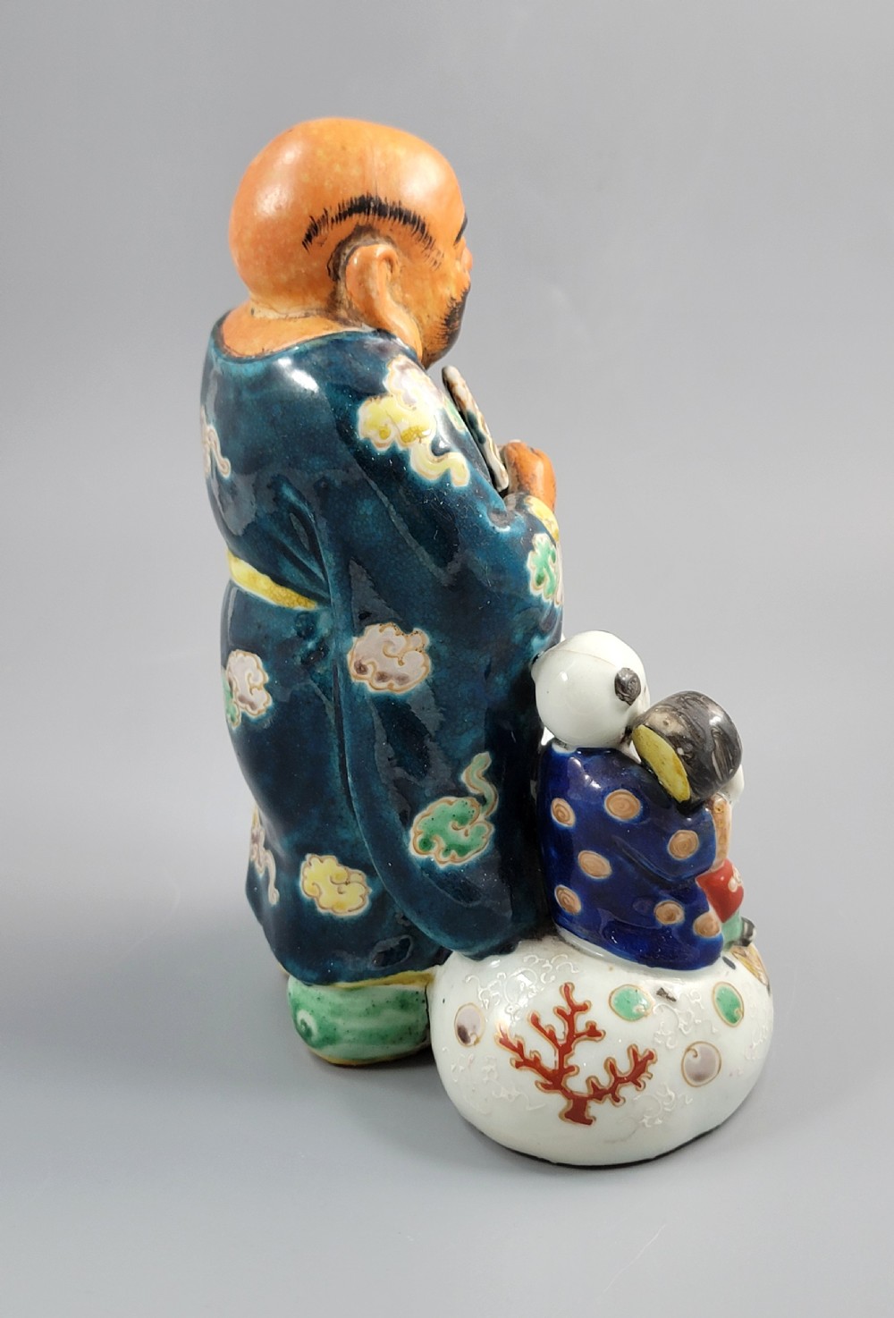 A Japanese Satsuma earthenware figure depicting Hotei in patterned blue robes holding a fan, standing next to a small child on a patterned sack. View VI