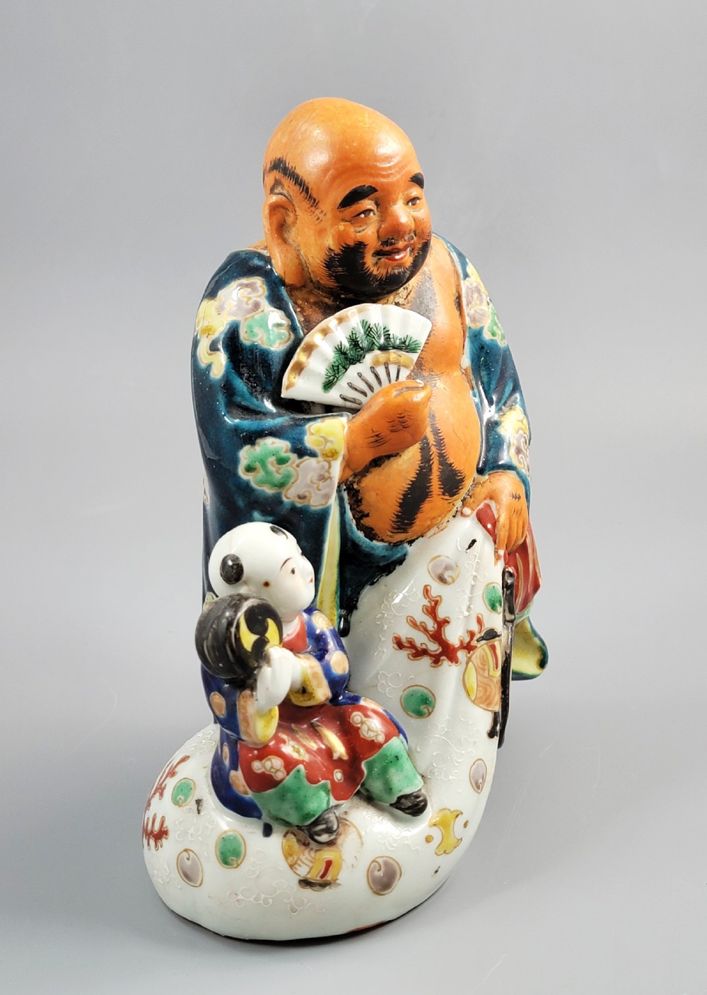 A Japanese Satsuma earthenware figure depicting Hotei in patterned blue robes holding a fan, standing next to a small child on a patterned sack. View VII