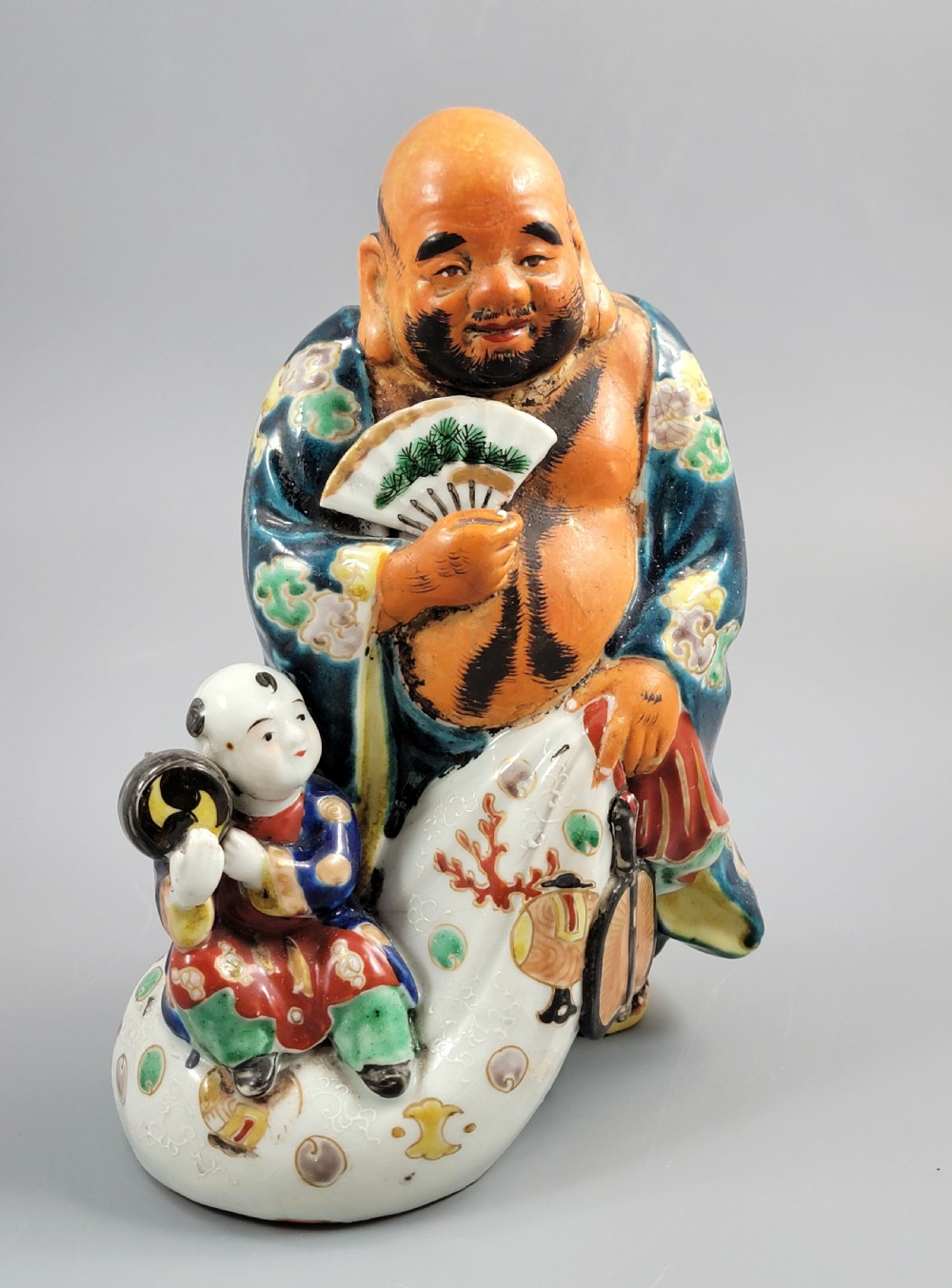 A Japanese Satsuma earthenware figure depicting Hotei in patterned blue robes holding a fan, standing next to a small child on a patterned sack. View VIII