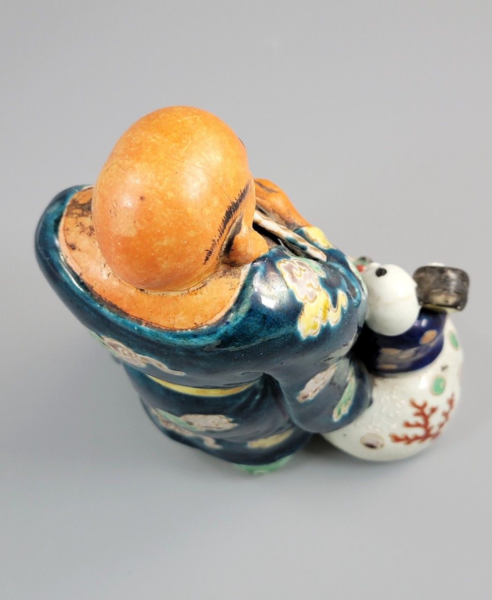 A Japanese Satsuma earthenware figure depicting Hotei in patterned blue robes holding a fan, standing next to a small child on a patterned sack. View IX
