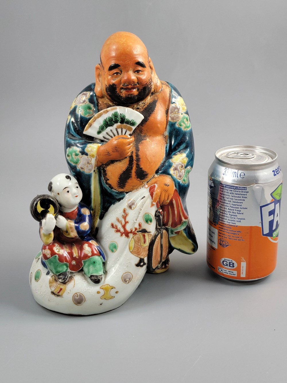 A Japanese Satsuma earthenware figure depicting Hotei in patterned blue robes holding a fan, standing next to a small child on a patterned sack. View XIV