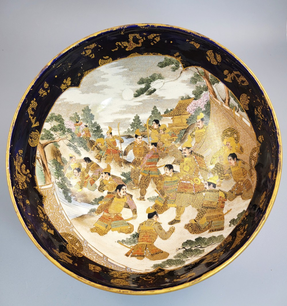 A circular Japanese Satsuma earthenware bowl featuring a central scene of samurai warriors in gold and polychrome enamels surrounded by a dark cobalt blue rim decorated with intricate gold scrollwork.