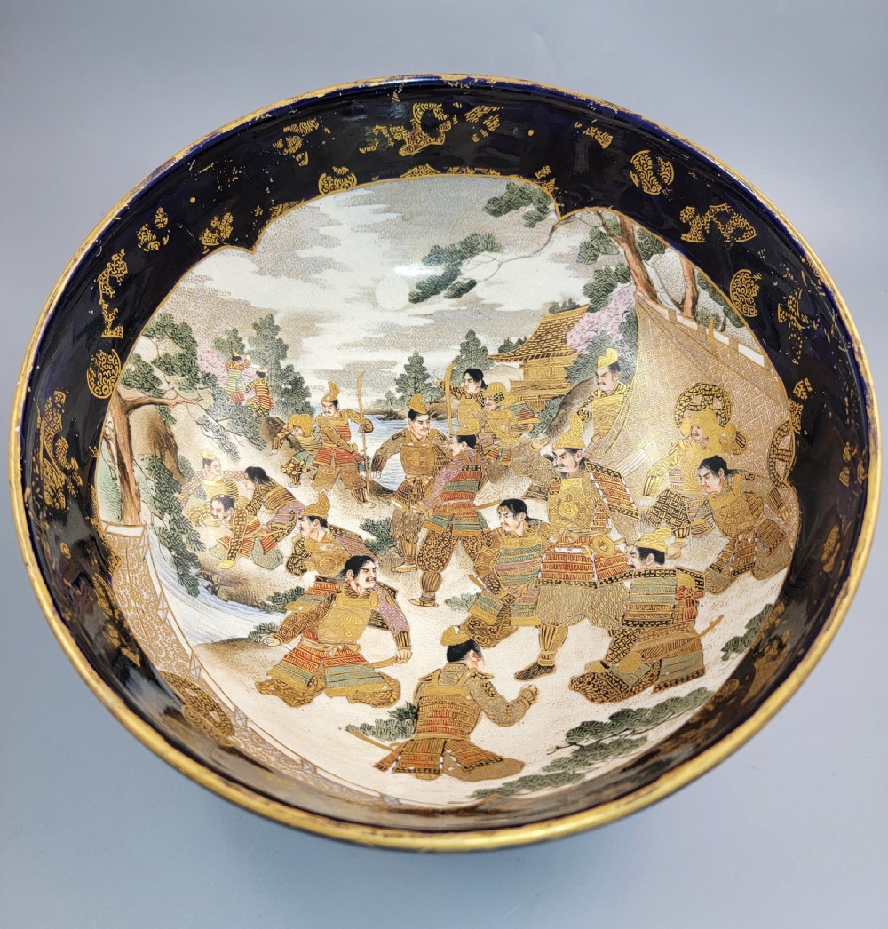 A circular Japanese Satsuma earthenware bowl featuring a central scene of samurai warriors in gold and polychrome enamels surrounded by a dark cobalt blue rim decorated with intricate gold scrollwork. View III