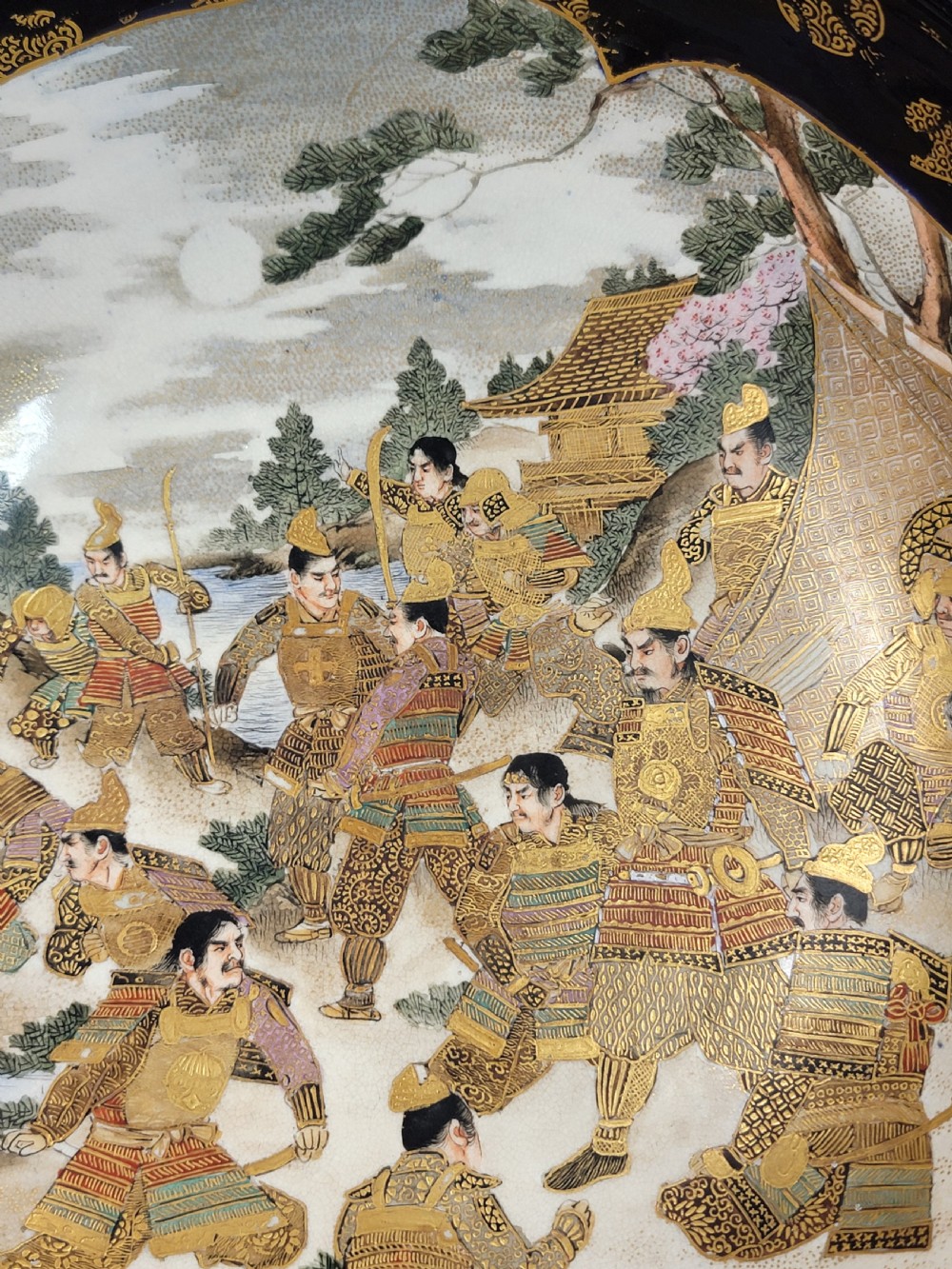 A circular Japanese Satsuma earthenware bowl featuring a central scene of samurai warriors in gold and polychrome enamels surrounded by a dark cobalt blue rim decorated with intricate gold scrollwork. View IV