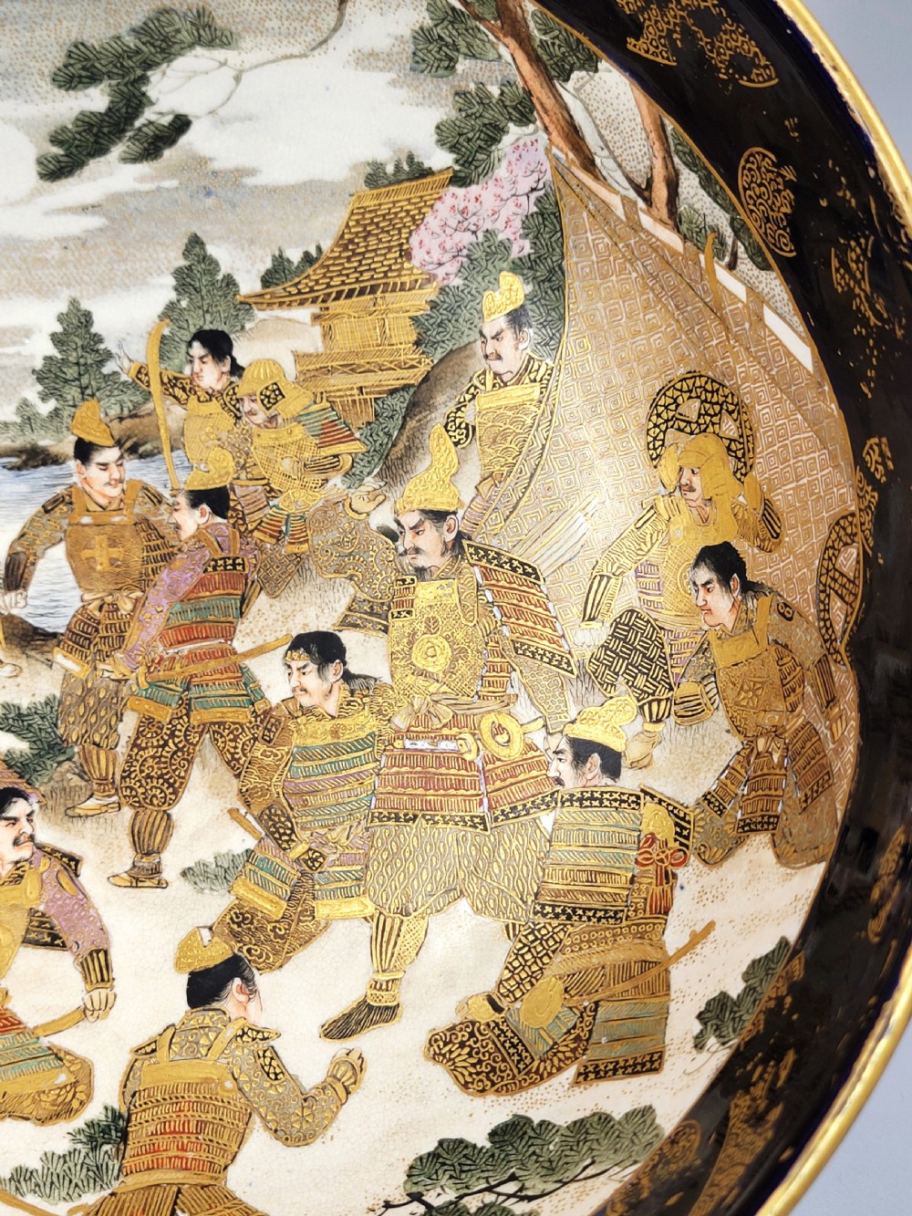 A circular Japanese Satsuma earthenware bowl featuring a central scene of samurai warriors in gold and polychrome enamels surrounded by a dark cobalt blue rim decorated with intricate gold scrollwork. View V