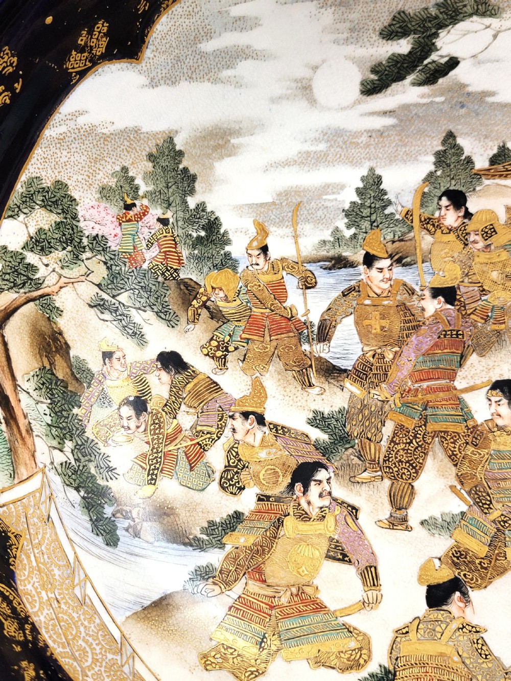 A circular Japanese Satsuma earthenware bowl featuring a central scene of samurai warriors in gold and polychrome enamels surrounded by a dark cobalt blue rim decorated with intricate gold scrollwork. View VI