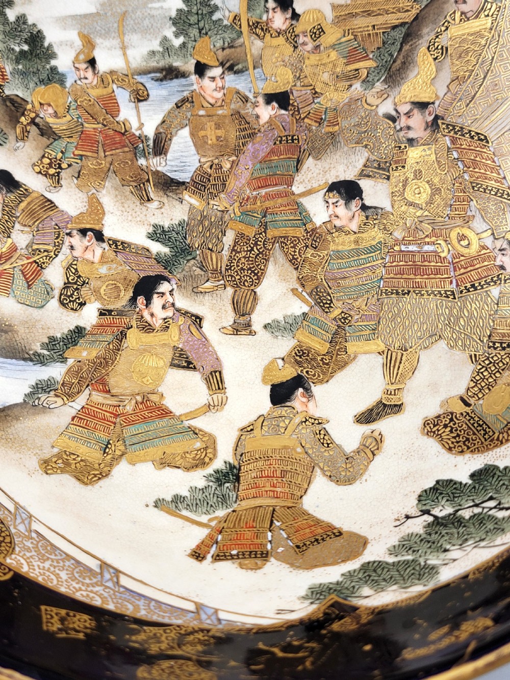 A circular Japanese Satsuma earthenware bowl featuring a central scene of samurai warriors in gold and polychrome enamels surrounded by a dark cobalt blue rim decorated with intricate gold scrollwork. View VII