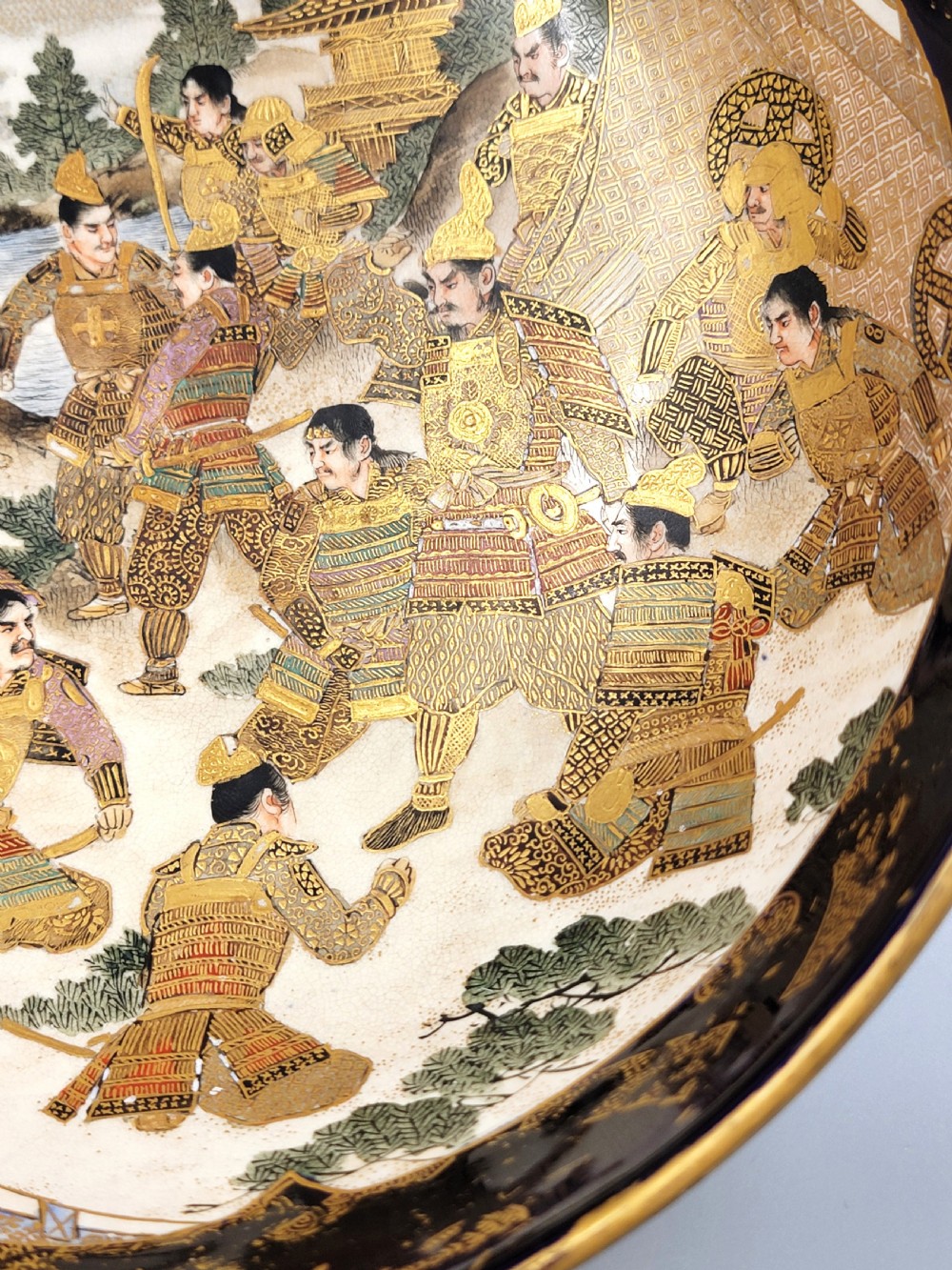 A circular Japanese Satsuma earthenware bowl featuring a central scene of samurai warriors in gold and polychrome enamels surrounded by a dark cobalt blue rim decorated with intricate gold scrollwork. View VIII