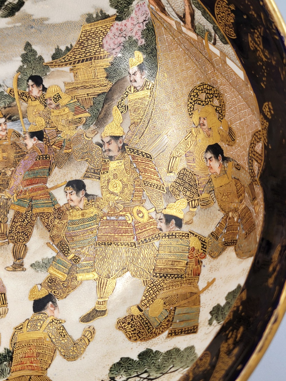 A circular Japanese Satsuma earthenware bowl featuring a central scene of samurai warriors in gold and polychrome enamels surrounded by a dark cobalt blue rim decorated with intricate gold scrollwork. View IX
