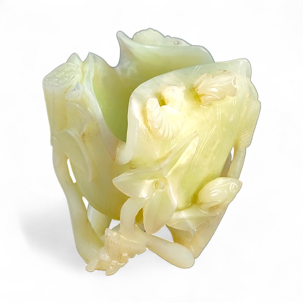 A pale celadon green jade cup sculpted into the form of a lotus leaf with surrounding stems and buds.