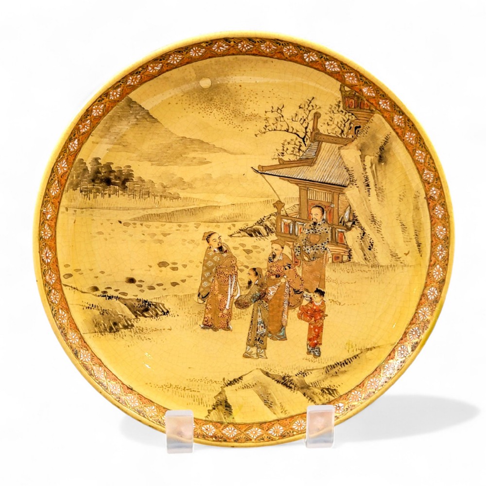 A circular Japanese Satsuma earthenware plate featuring a fine crackle glaze, detailed polychrome figures in traditional dress near a landscape building, and a gilded geometric border.