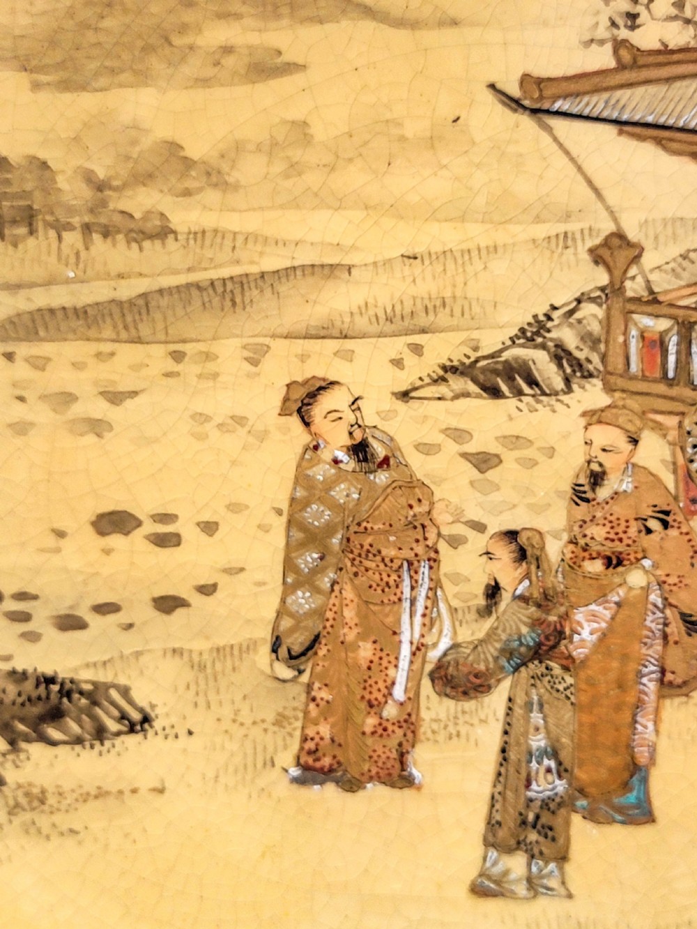 A circular Japanese Satsuma earthenware plate featuring a fine crackle glaze, detailed polychrome figures in traditional dress near a landscape building, and a gilded geometric border. View IV