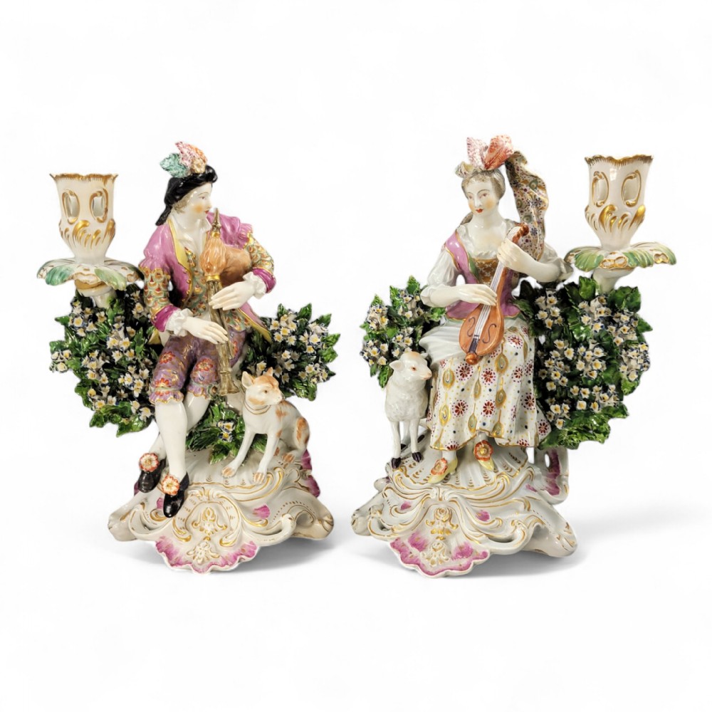 A pair of 18th-century Derby porcelain candlestick figures depicting a male bagpipe player with a dog and a female mandolin player with a lamb, set against floral boscage backgrounds on gilded scrollwork bases.