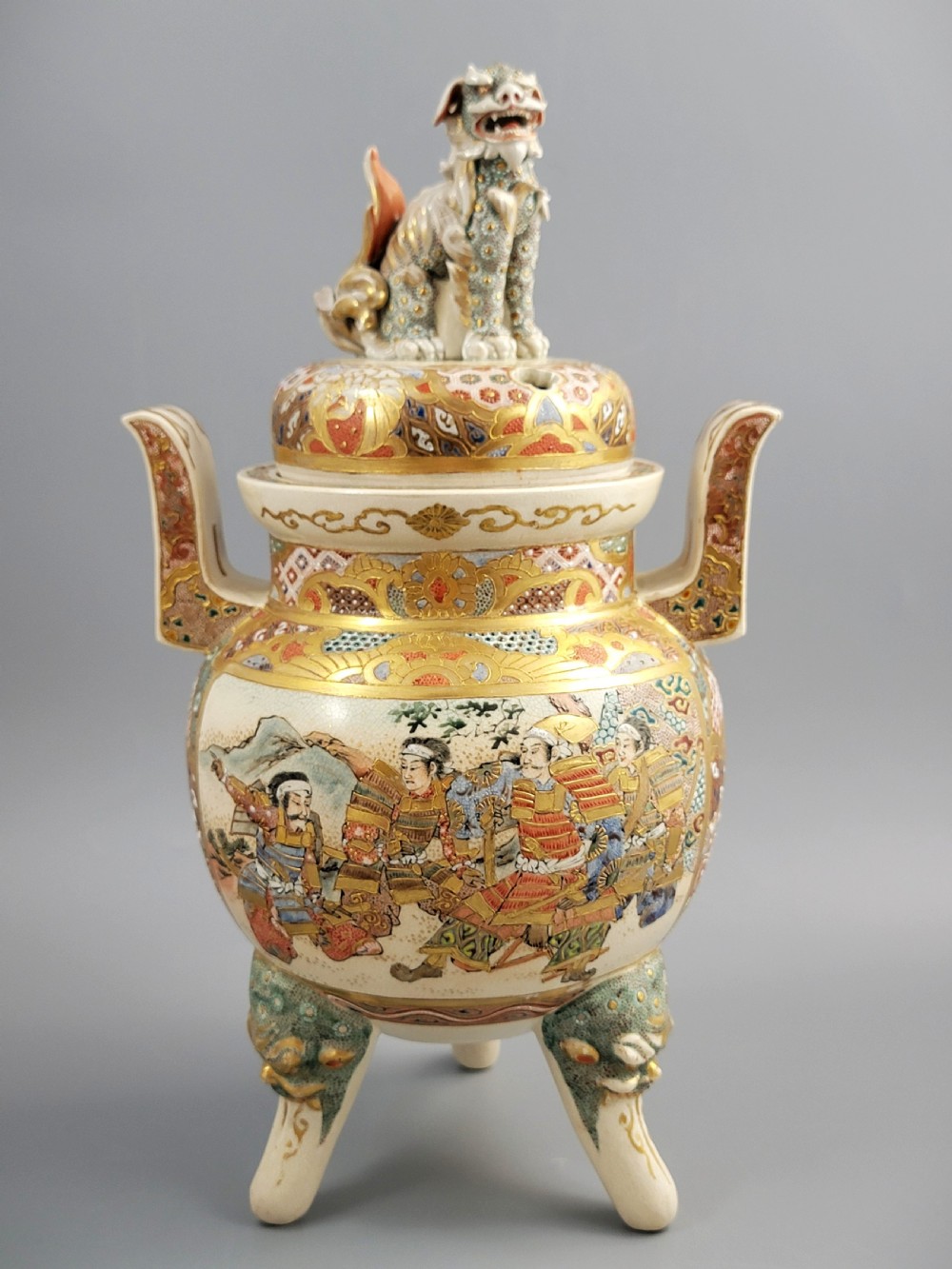 A cream-coloured Japanese Satsuma earthenware incense burner featuring a lid with a shishi lion finial, upright handles, tripod feet, and detailed polychrome paintings of samurai warriors with gold accents.