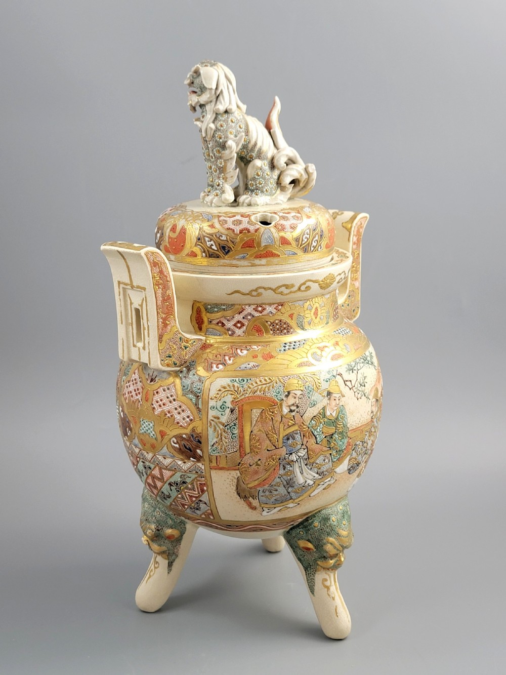 A cream-coloured Japanese Satsuma earthenware incense burner featuring a lid with a shishi lion finial, upright handles, tripod feet, and detailed polychrome paintings of samurai warriors with gold accents. View IV