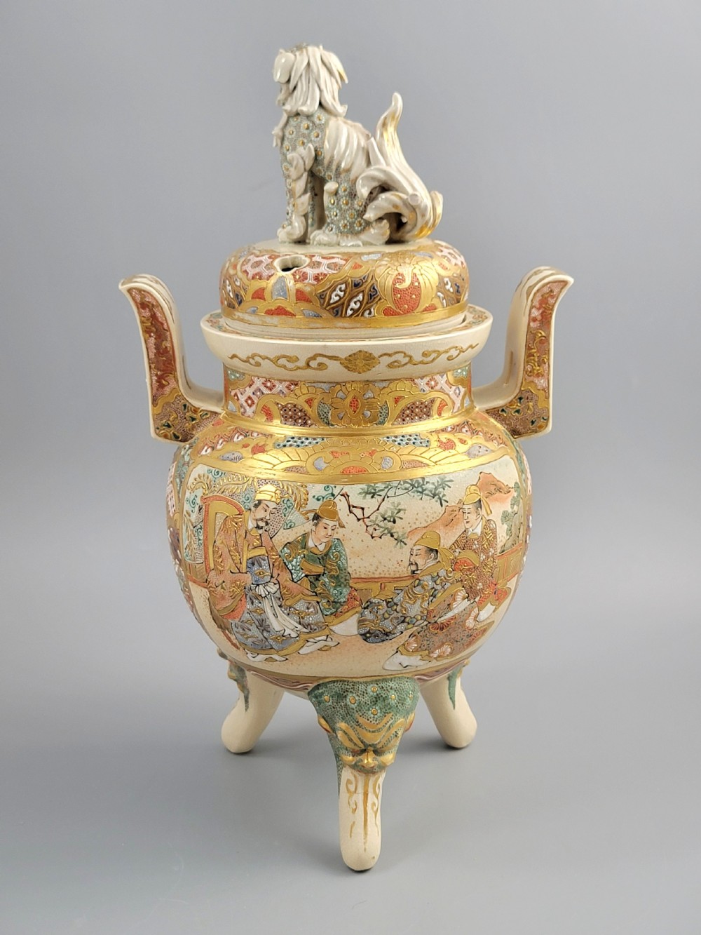 A cream-coloured Japanese Satsuma earthenware incense burner featuring a lid with a shishi lion finial, upright handles, tripod feet, and detailed polychrome paintings of samurai warriors with gold accents. View V