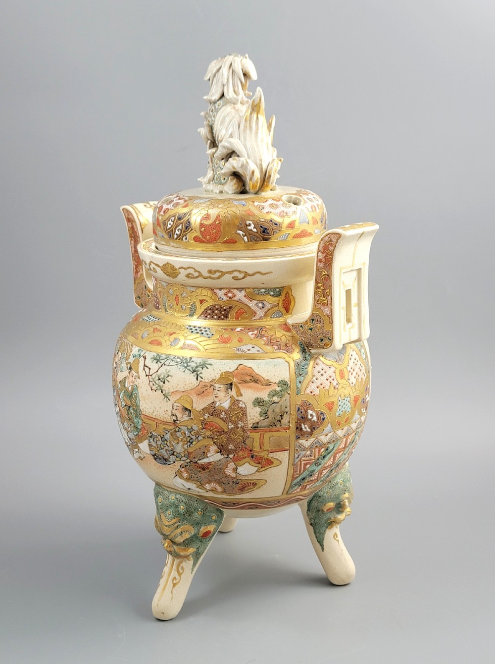 A cream-coloured Japanese Satsuma earthenware incense burner featuring a lid with a shishi lion finial, upright handles, tripod feet, and detailed polychrome paintings of samurai warriors with gold accents. View VI