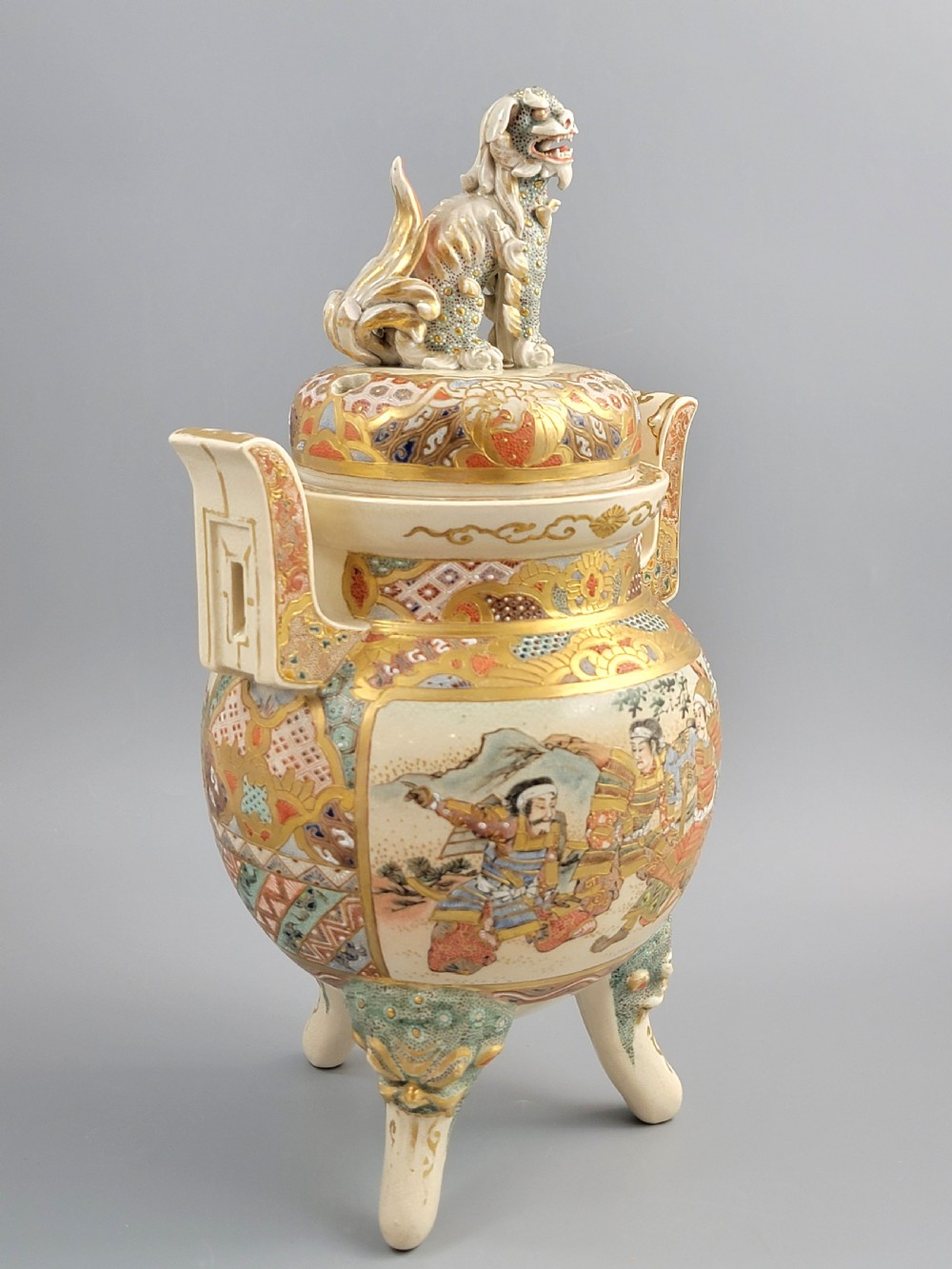 A cream-coloured Japanese Satsuma earthenware incense burner featuring a lid with a shishi lion finial, upright handles, tripod feet, and detailed polychrome paintings of samurai warriors with gold accents. View VIII