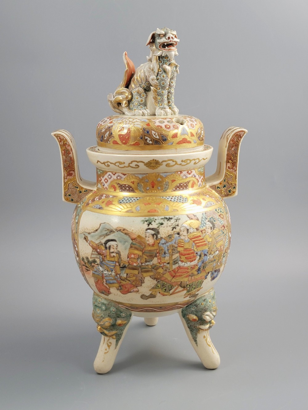 A cream-coloured Japanese Satsuma earthenware incense burner featuring a lid with a shishi lion finial, upright handles, tripod feet, and detailed polychrome paintings of samurai warriors with gold accents. View IX