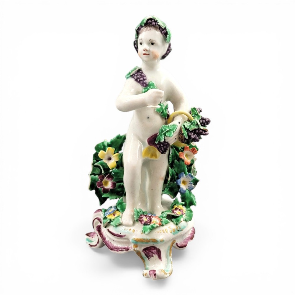 An 18th-century Bow soft-paste porcelain figure of a putto adorned with grapevines, standing on a Rococo scroll base with colourful floral bocage behind.