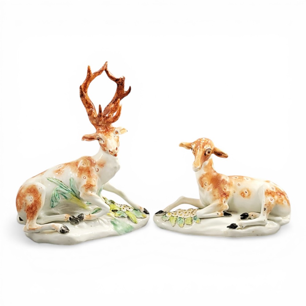 A pair of 18th-century Derby porcelain figures depicting a reclining stag with branched antlers and a doe, painted with chestnut-spotted coats on floral-adorned white bases.
