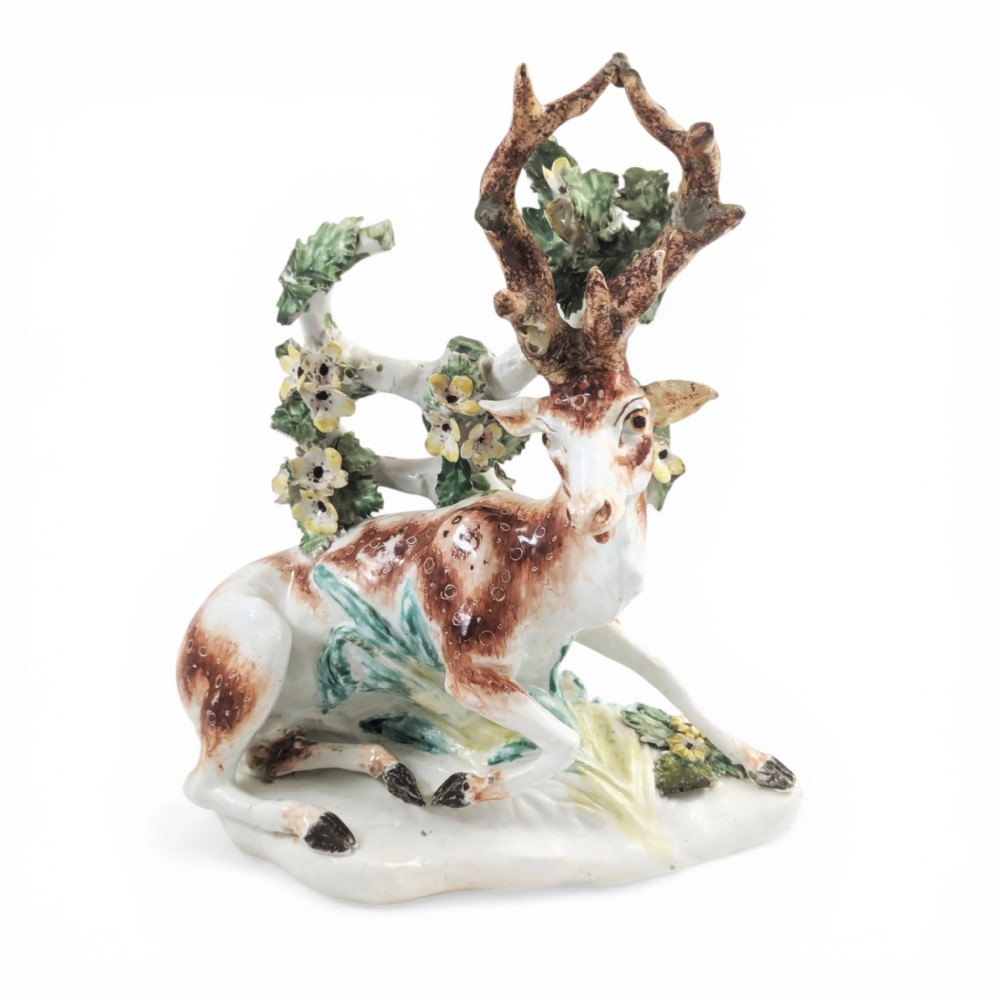 An 18th-century soft-paste porcelain figurine depicting a recumbent brown and white spotted stag resting in front of a leafy bocage with small yellow flowers.