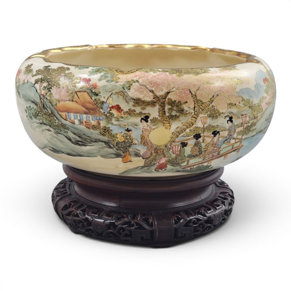 A Japanese Satsuma earthenware bowl with hand-painted figures under trees and gold accents, resting on a dark carved wooden stand.