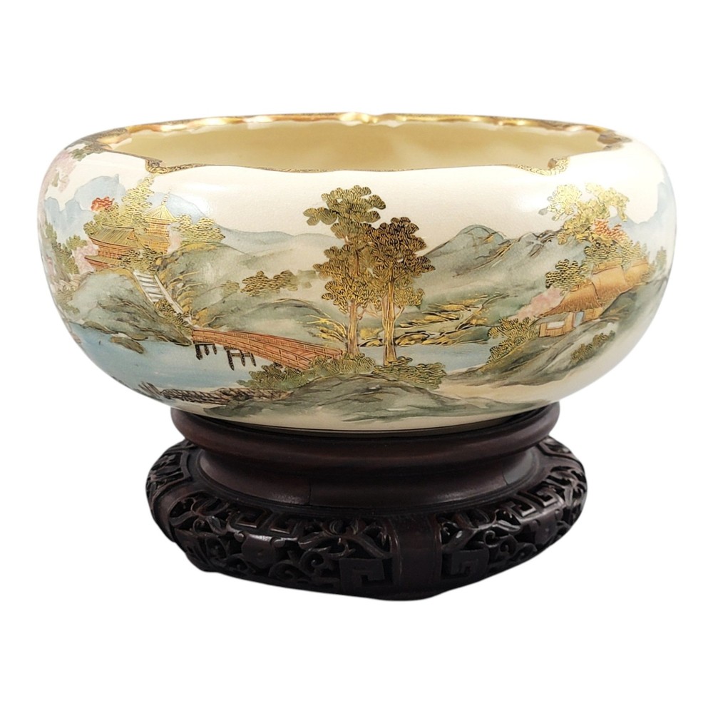 A Japanese Satsuma earthenware bowl with hand-painted figures under trees and gold accents, resting on a dark carved wooden stand. View VIII