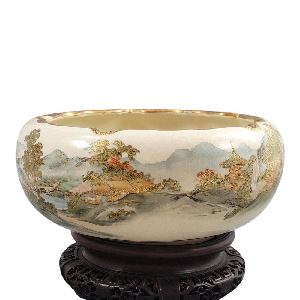 A Japanese Satsuma earthenware bowl with hand-painted figures under trees and gold accents, resting on a dark carved wooden stand. View IX
