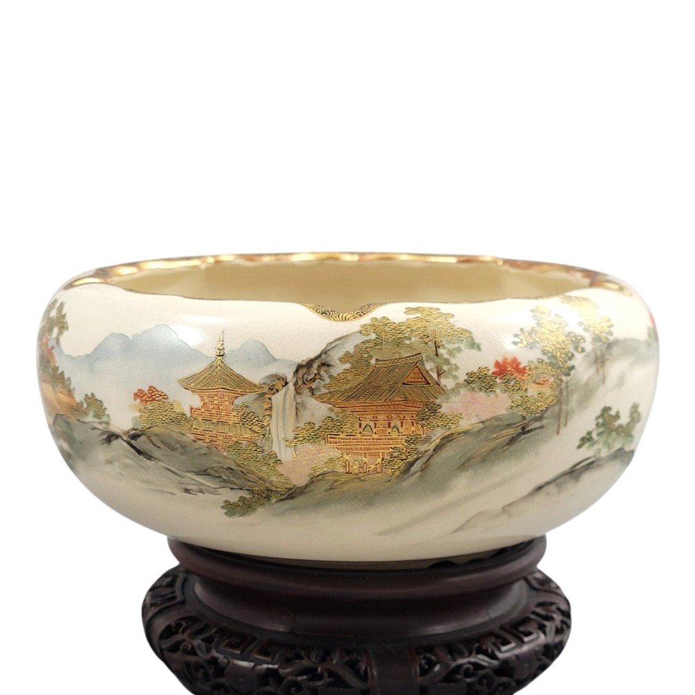 A Japanese Satsuma earthenware bowl with hand-painted figures under trees and gold accents, resting on a dark carved wooden stand. View X